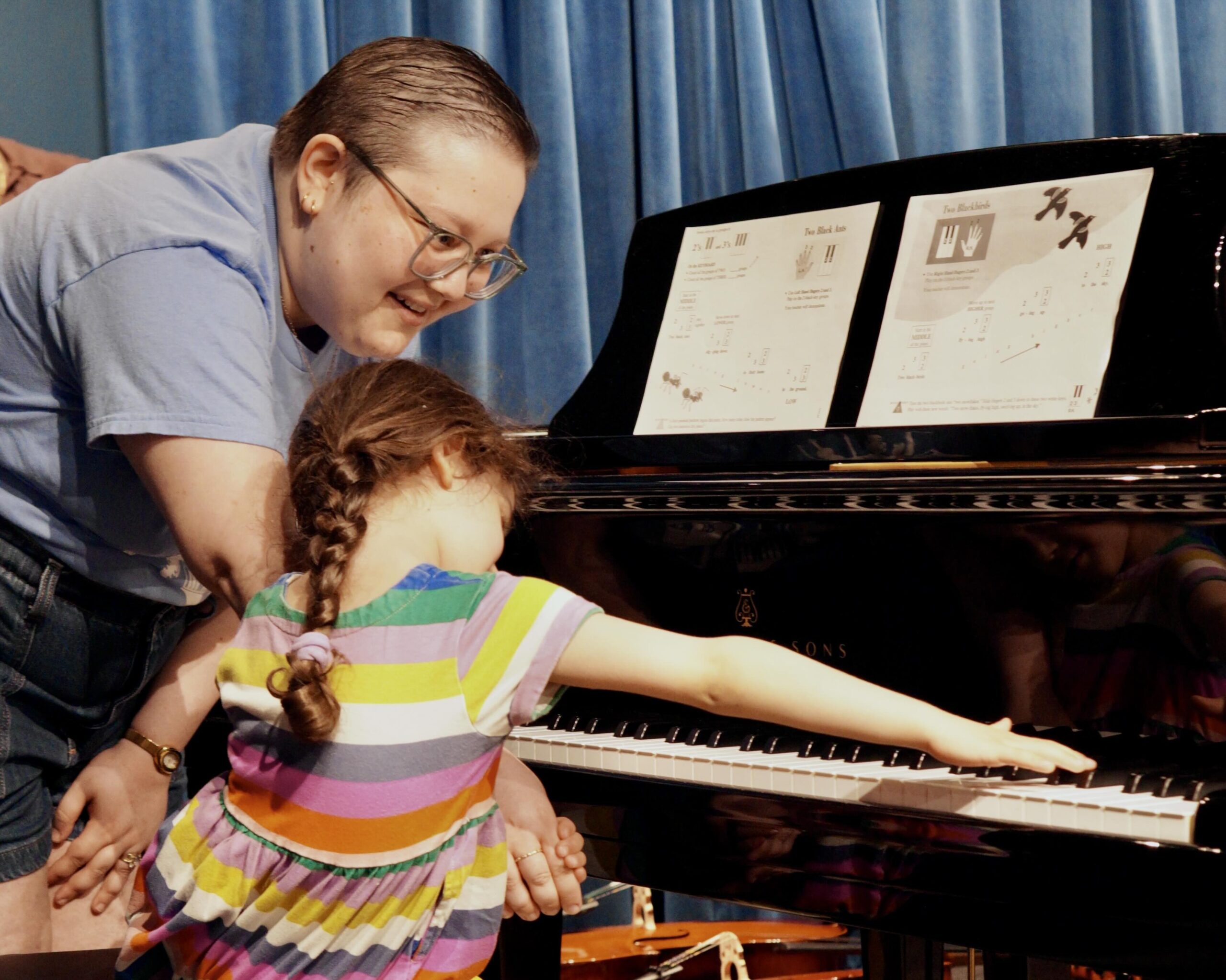 A young girl in a rainbow-striped dress reaches across the keys of a grand piano as a Bloomingdale teacher guides her