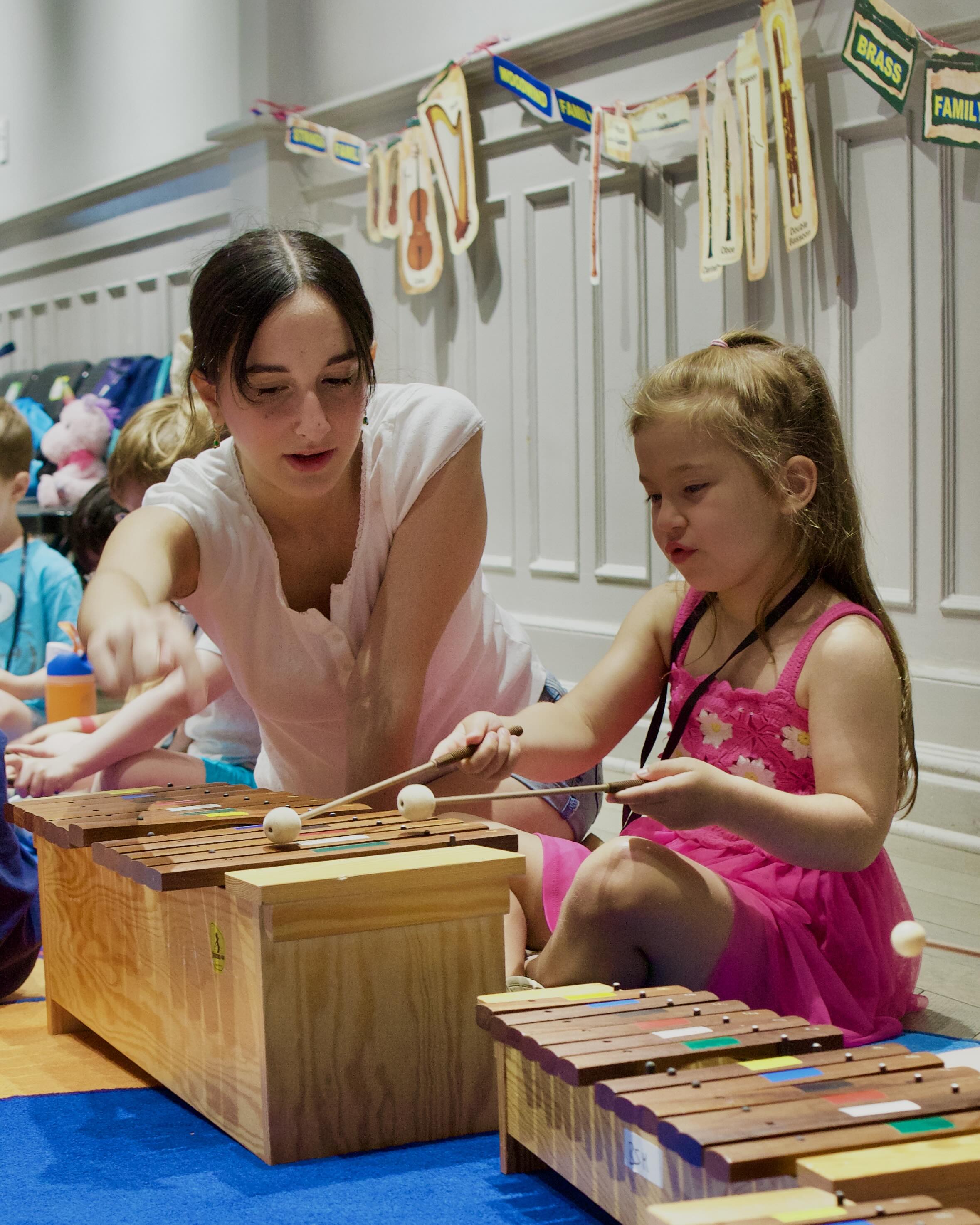 A young girl plays a color-coded xylophone with mallets while a teacher points to the keys during Meet the Instruments at Bloomingdale School of Music