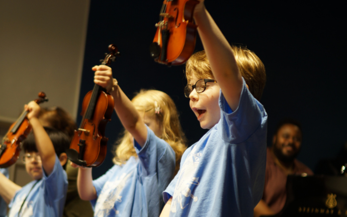 A boy in Meet the instruments, early childhood class, holding a violin in the air with his friends.