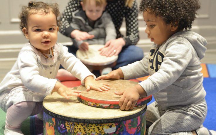 an image of babies around 1-2 years old playing the drum, smiling, and having a fun time with their caregiver or parent
